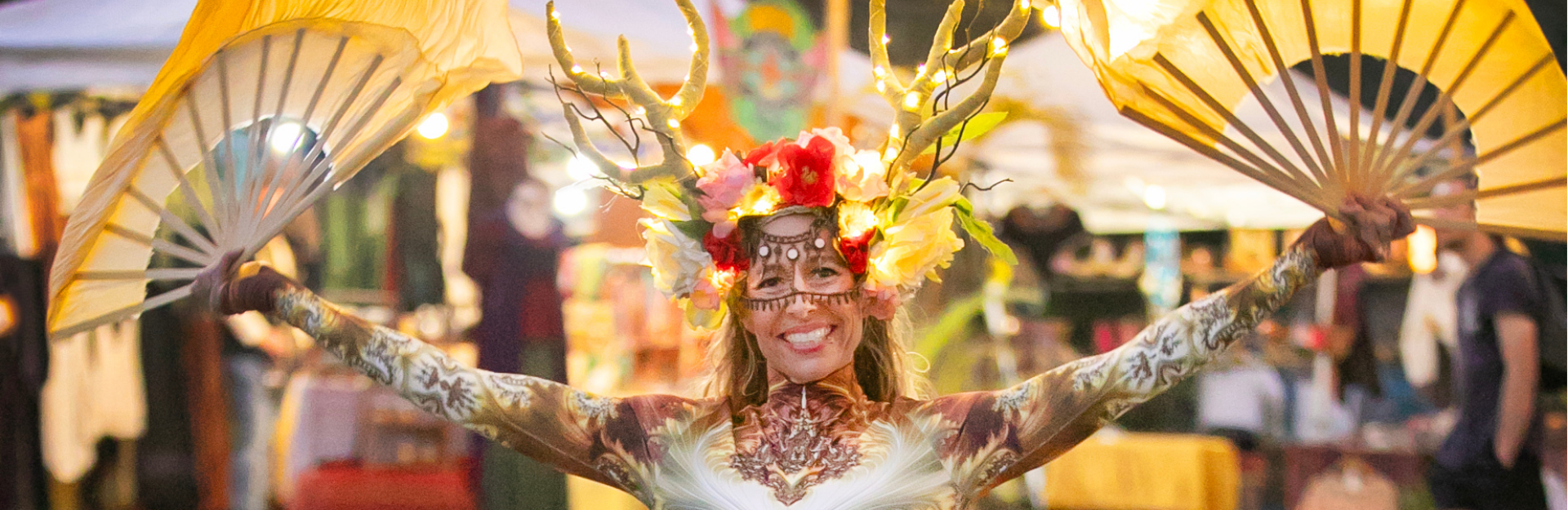 A photo from Pranafest at night, showing a costumed performer with glowing fans posing in a lit festival walkway