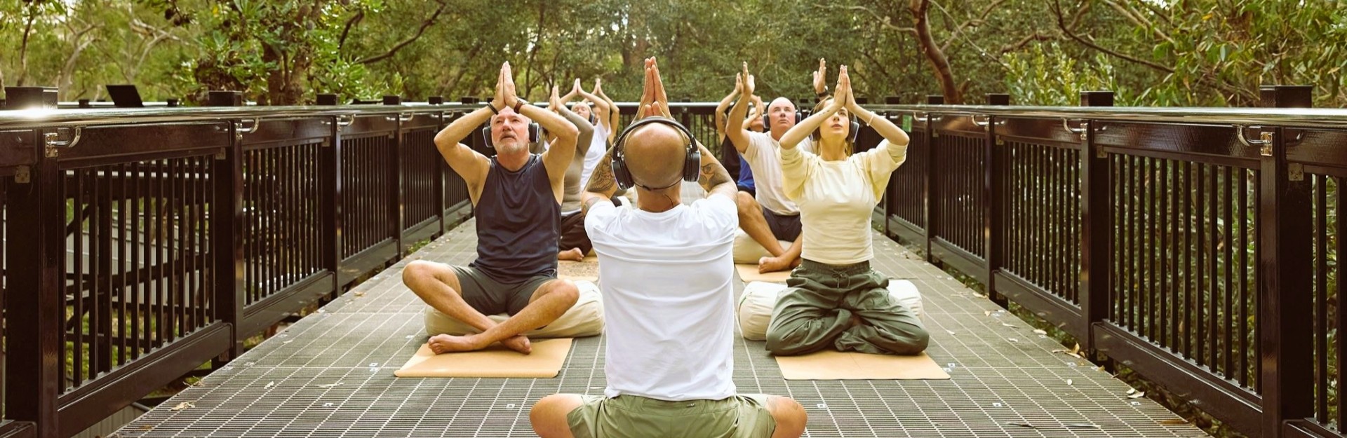 a group of people sitting on an elevated walkway among the gumtrees, wearing headphones and doing a meditation
