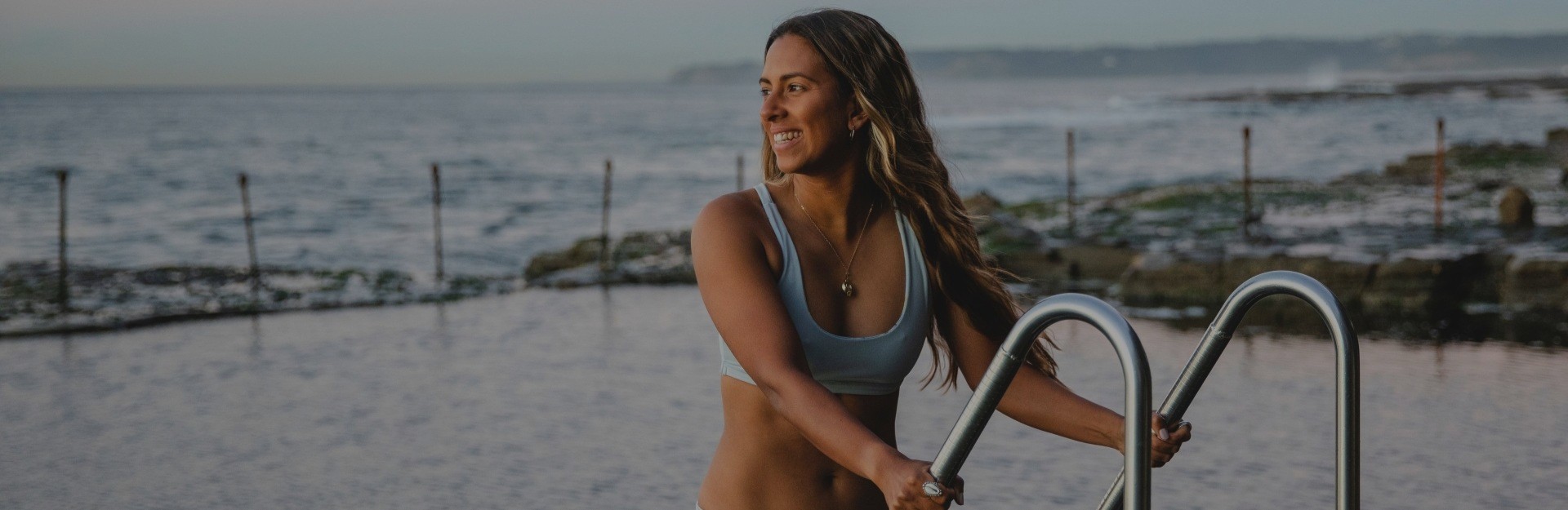 A young woman in a bikini climbing a ladder out of an ocean pool at sunrise