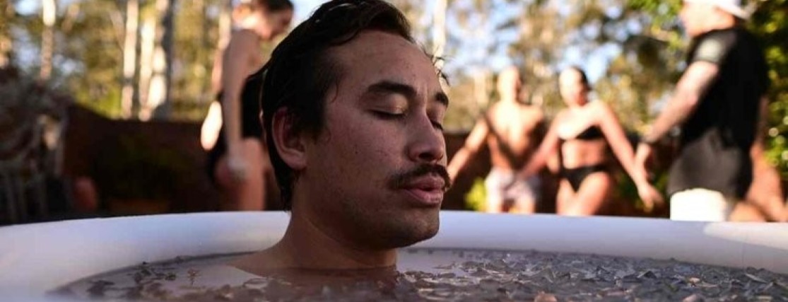 a young man immersed in a barrel of ice water up to his shoulders with his eyes closed