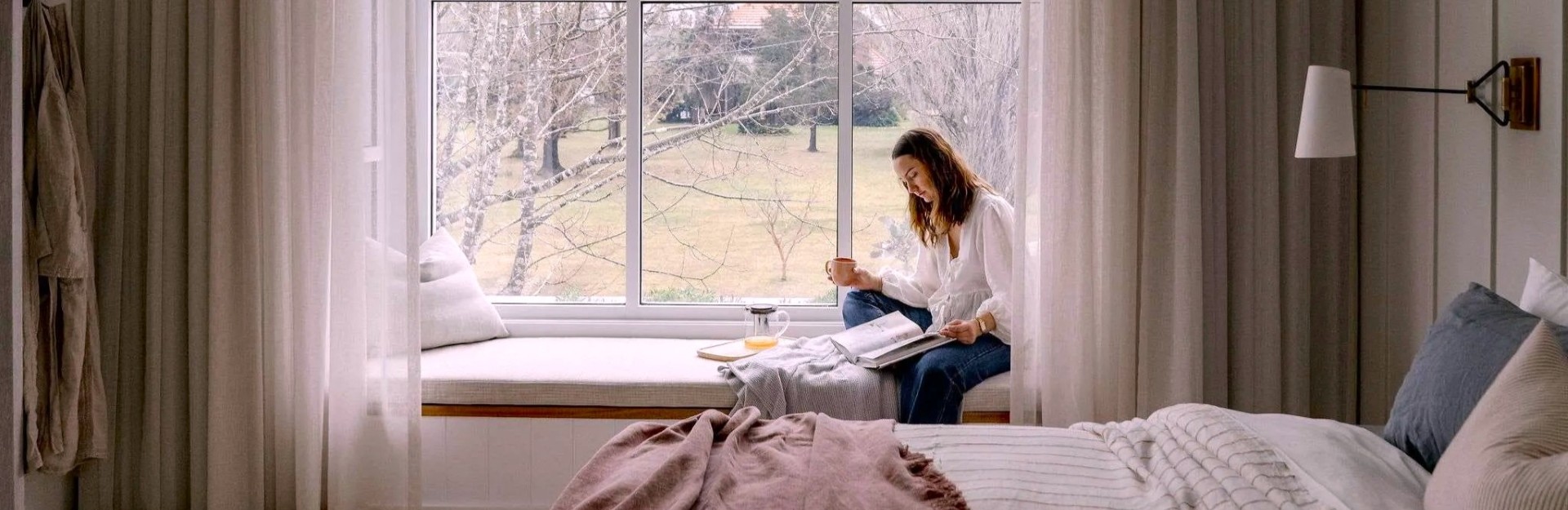 A lady reading with a cup of tea next to a window in a beautiful bedroom with a king bed and pastel soft furnishings