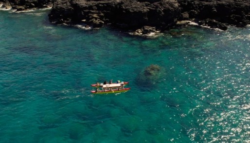 An aerial image of people in a traditional Hawaiian double hulled canoe, paddling along the Kona Coast