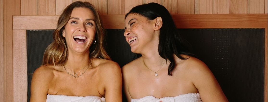 2 ladies wearing white towels laughing in a sauna