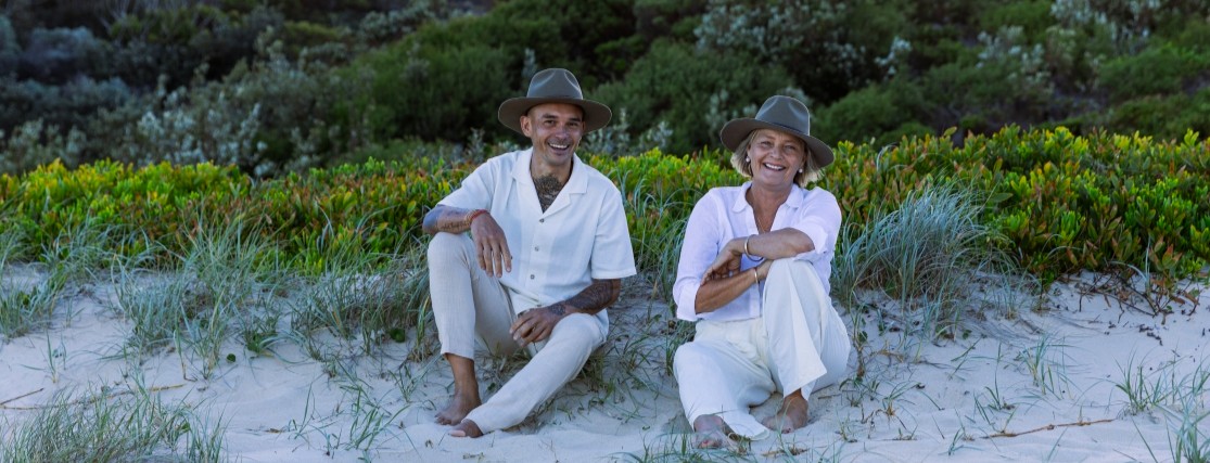 a man and a woman dressed in white sitting on the sand in front of green vegetation