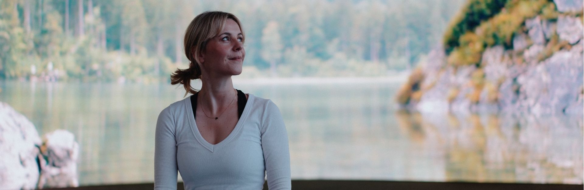 A yoga teacher wearing a long sleeve white top, sitting on the floor in front of a photo of a lake in a forest