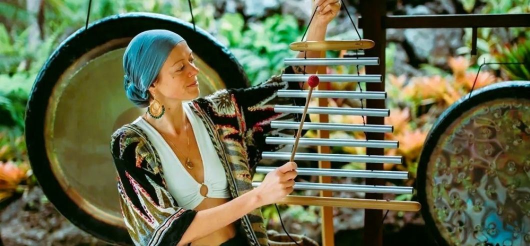 A lady kneeling on the ground in front of a set of gongs, plating a musical instrument in a sound healing session