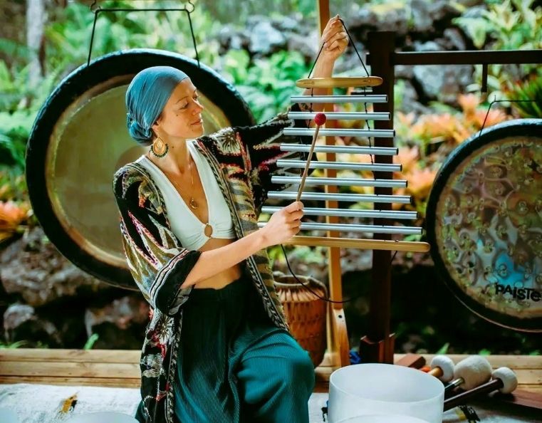 A lady kneeling on the ground in front of a set of gongs, plating a musical instrument in a sound healing session