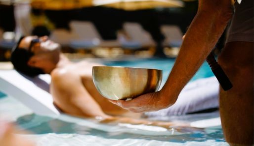 a man lying on a sunbed in a pool, with someone hosting a sound bath with a metal bowl