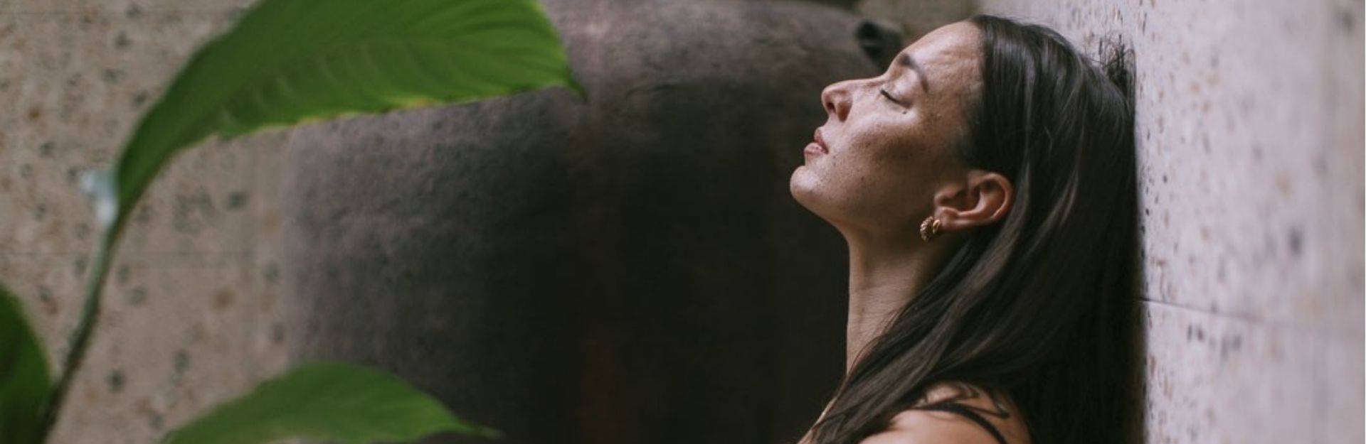 a dark-haired lady in a black swimsuit resting against a wall with her eyes closed