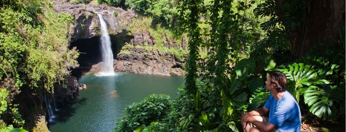 A man sitting in the forest looking towards Rainbow Falls on the Island of hawaii