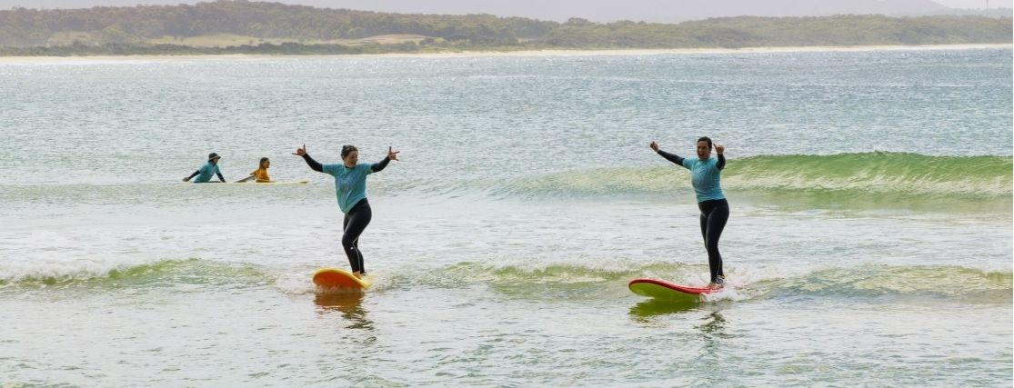 2 women surfing with their hands in the air, giving the thumbs up