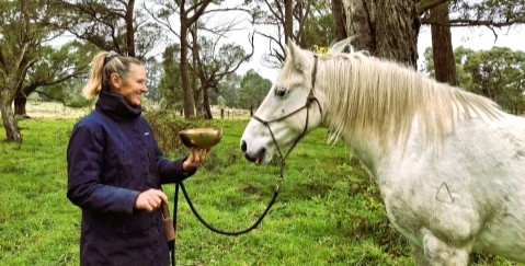 A woman standing in front of a white horse, holding a singing bowl