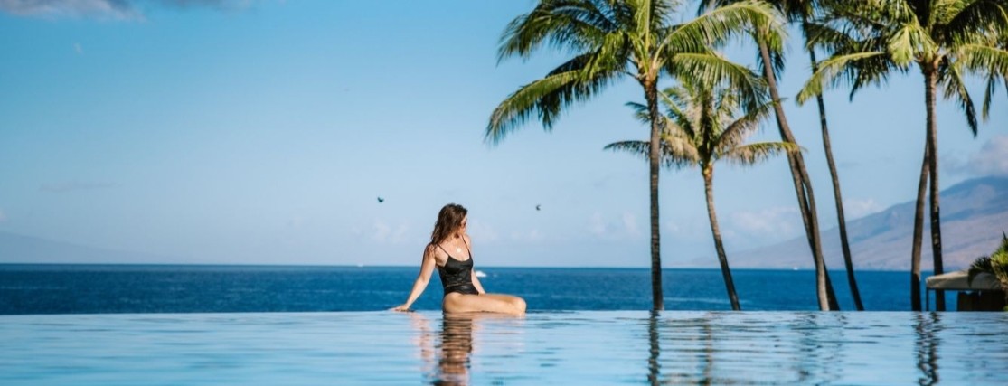 a lady in a black swimsuit sitting on the edge of an infinity pool near the ocean in Maui