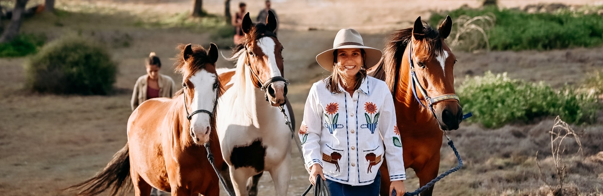 A lady in a hat leading 3 brown horses along a trail on a property