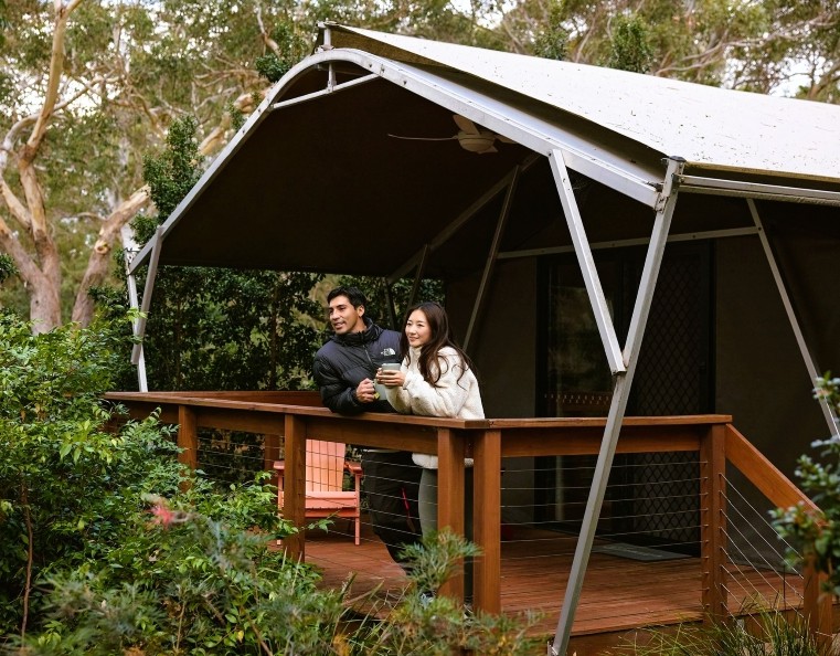 A young couple standing outside a glamping tent in the Australian bush with a cup of tea