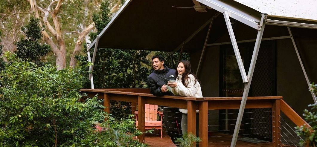 A young couple standing outside a glamping tent in the Australian bush with a cup of tea