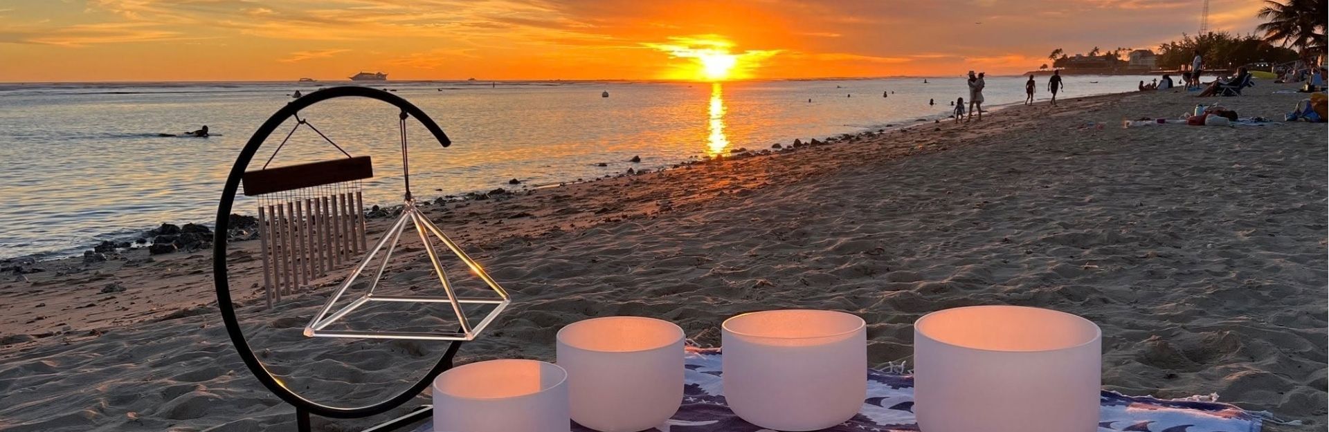 a sound healing session set up at sunset on the beach with white crystal bowls laied out in a semi circle and a prism hanging from a pedestal
