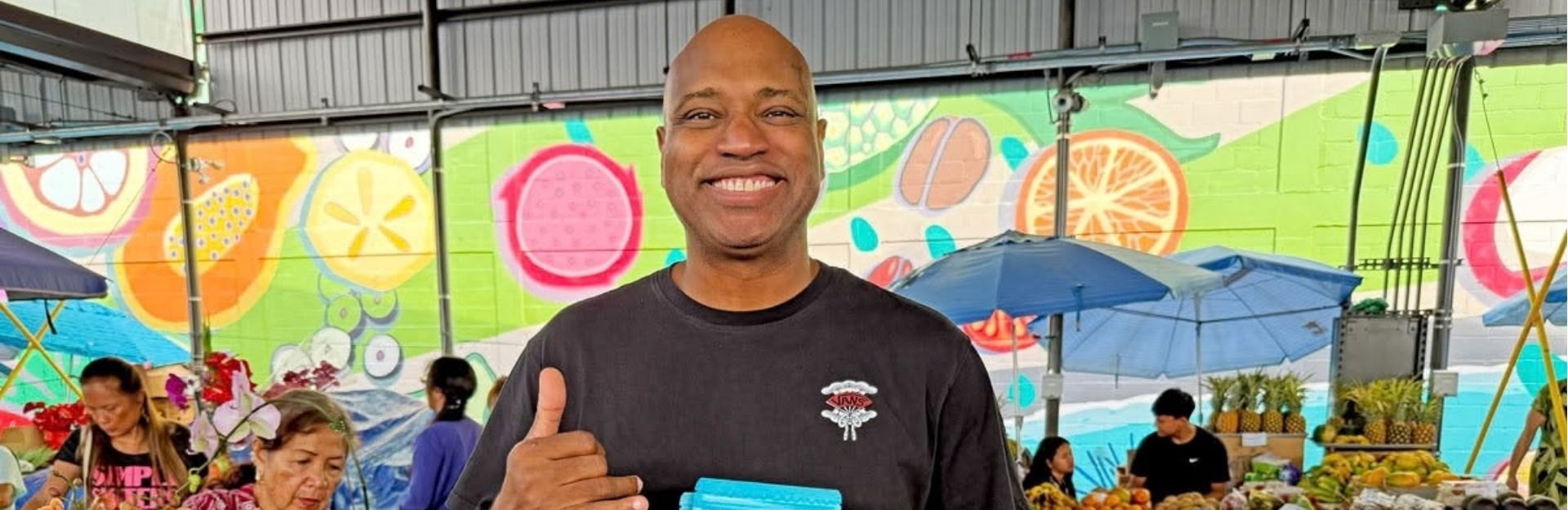 a man wearing a black t-shirt and doing the 'hang loose' sign while holding produce at a farmers market